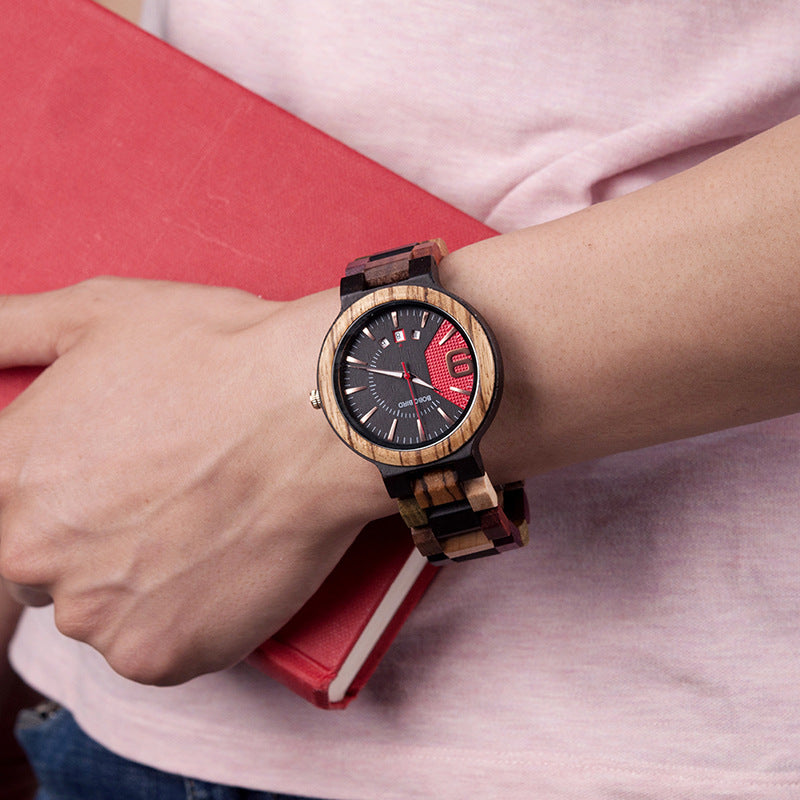 Men's wristwatch featuring a wooden watch face and colorful band, worn while holding a red notebook.